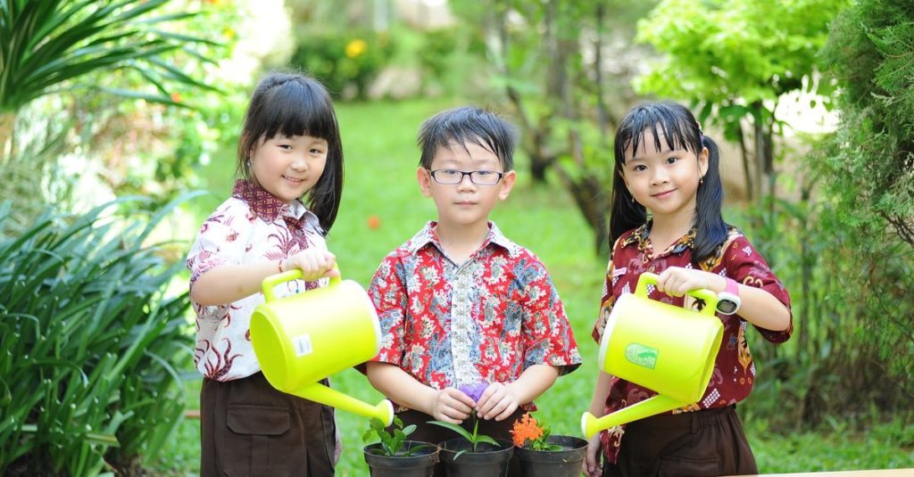 Tiga anak sedang menikmati berkebun di kebun hijau subur dengan penyiram tanaman.