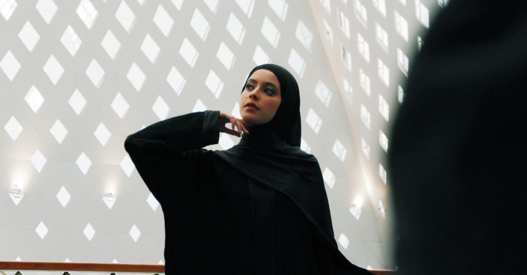 Seorang wanita yang berpikir dalam hijab berdiri di dalam ruangan di depan dinding bermotif, menangkap keanggunan dan identitas budaya.