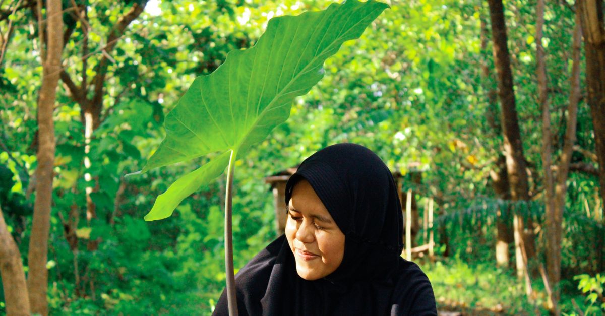 Seorang wanita mengenakan hijab tersenyum di antara pohon-pohon hijau subur, menikmati pemandangan luar ruangan.