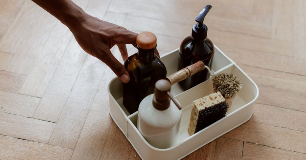 Sebuah tangan menjangkau set pembersihan dengan botol dan sikat di lantai kayu.