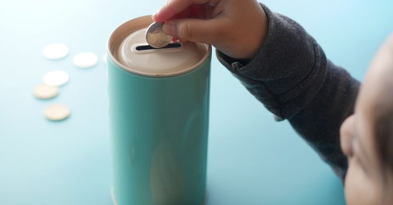 Seorang anak meletakkan koin ke dalam celengan berwarna teal, yang mewakili menabung dan pendidikan keuangan.