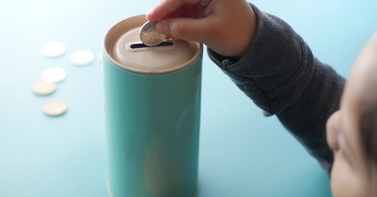 Seorang anak meletakkan koin ke dalam celengan berwarna teal, yang mewakili menabung dan pendidikan keuangan.