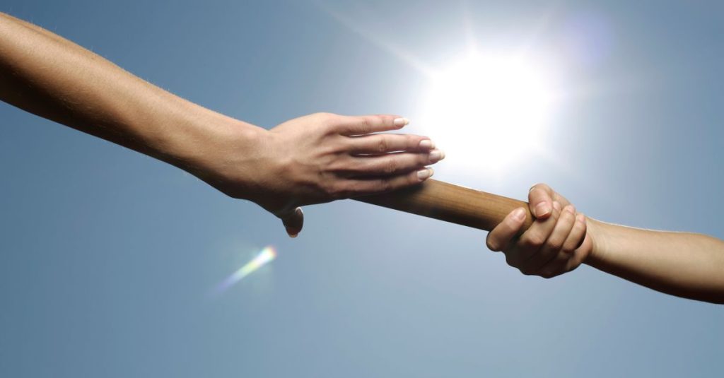 Dua tangan yang melewati baton di bawah sinar matahari yang cerah, melambangkan kerja sama dan kolaborasi.