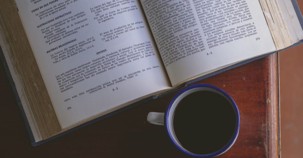 Pandangan dari atas sebuah buku terbuka dengan secangkir kopi di atas meja kayu dalam pencahayaan alami.
