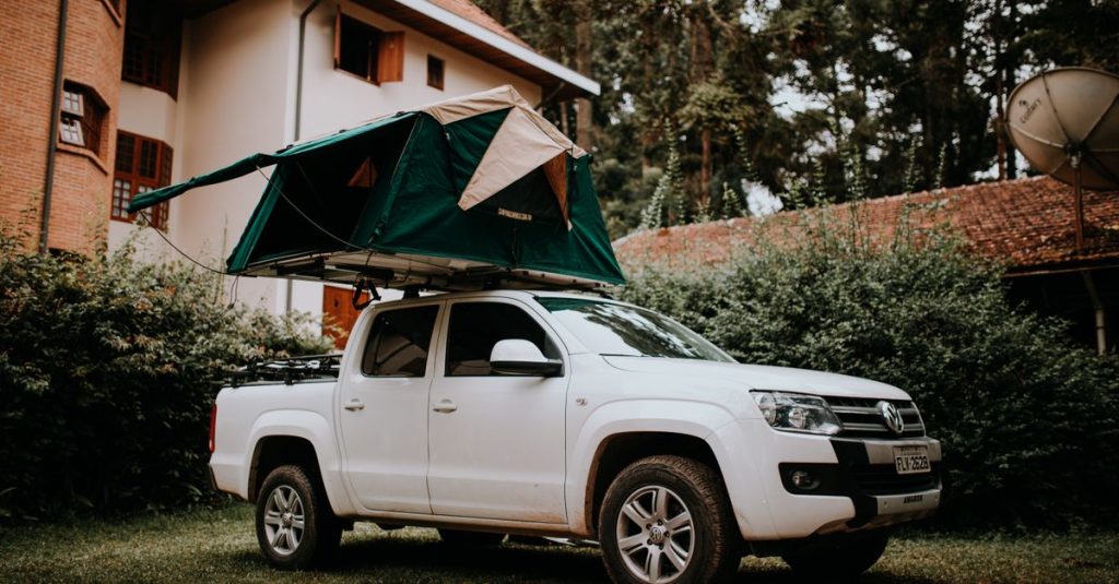 Truk pickup putih dengan tenda atap diparkir dekat rumah, ideal untuk tema camping dan perjalanan.