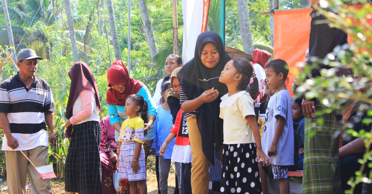 Pertemuan luar ruangan yang meriah dengan anak-anak dan orang dewasa berinteraksi di bawah sinar matahari dekat pohon.
