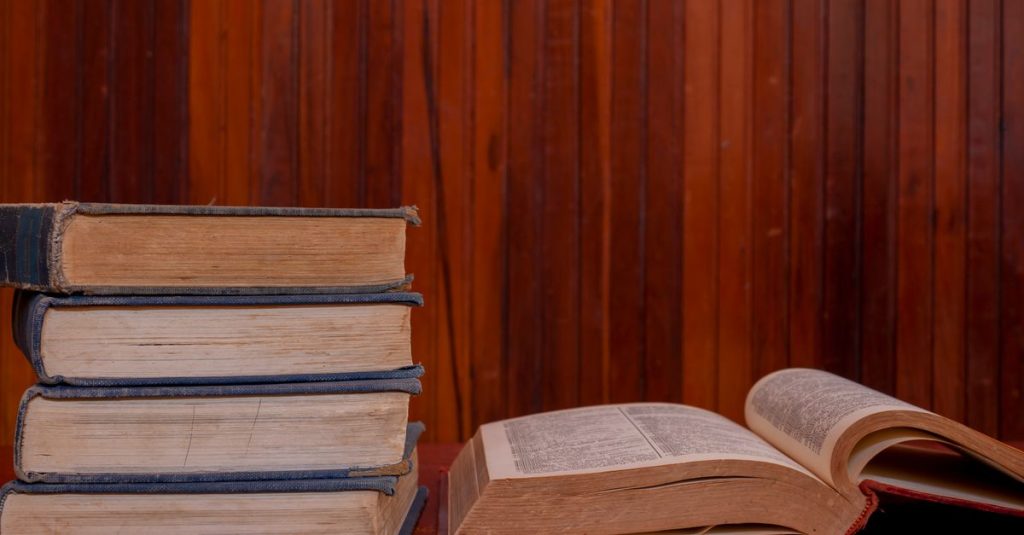 Sebuah tumpukan buku vintage dengan satu terbuka, diletakkan di atas meja kayu, melawan dinding berpanel kayu.
