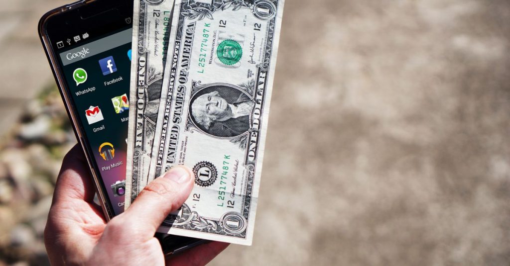Close-up tangan yang memegang uang kertas dolar AS dan smartphone di luar ruangan, menampilkan teknologi finansial.