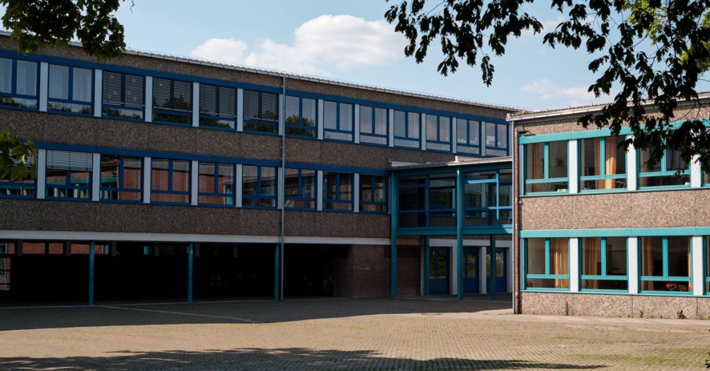 Tampilan luar bangunan pendidikan modern di Geilenkirchen, Jerman, yang memiliki jendela besar dan halaman yang dipaving.