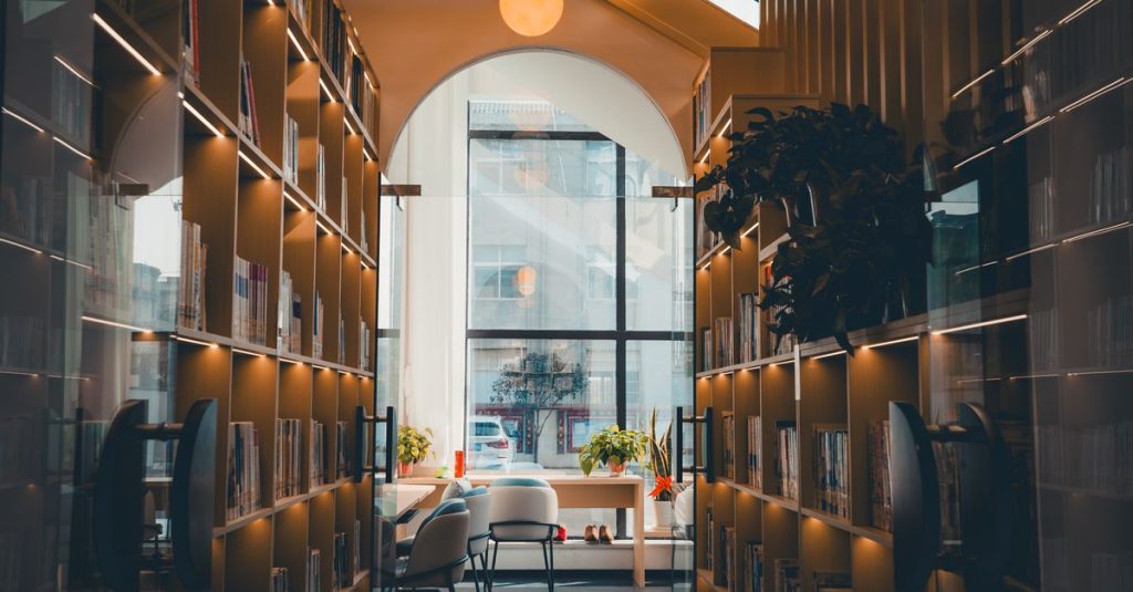 Sebuah interior perpustakaan yang hangat dan mengundang dengan rak buku, cahaya alami, dan dekorasi modern, ideal untuk membaca.