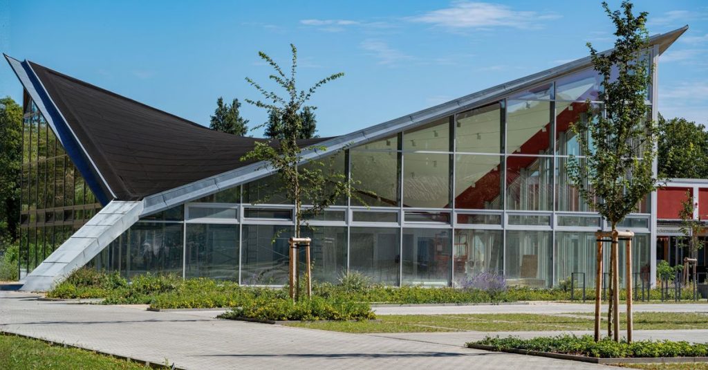 Sebuah gedung kaca modern yang mencolok dengan atap geometris tajam, di bawah langit biru yang cerah.