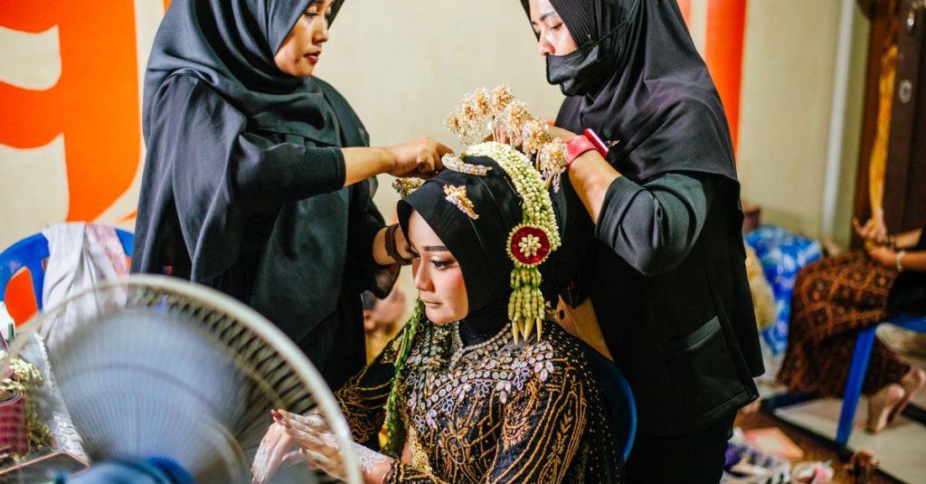 Dua perempuan membantu pengantin wanita dengan pakaian dan hiasan kepala tradisional Jawa di dalam ruangan.