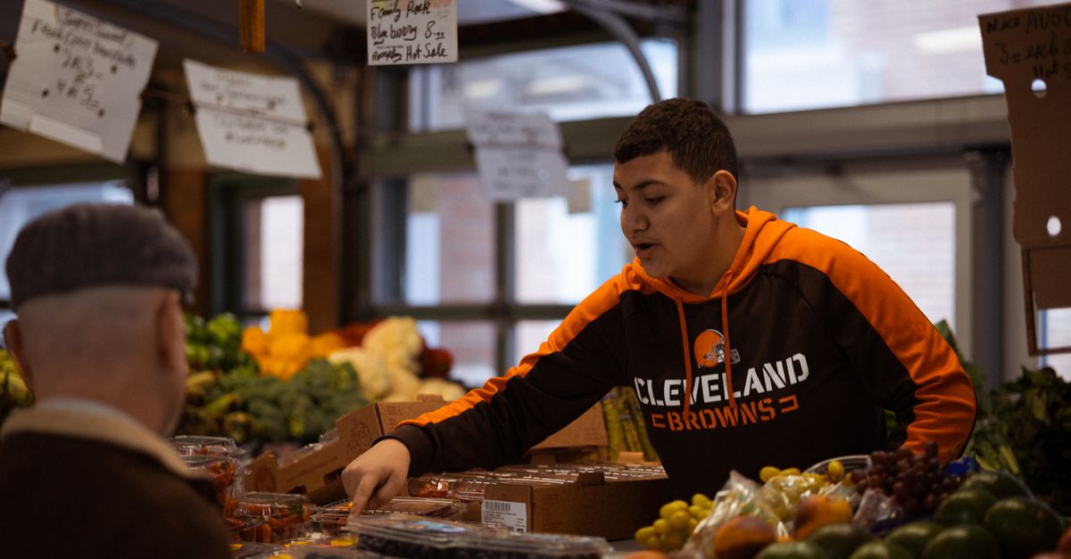 Seorang pria muda dengan hoodie Cleveland Browns menjual produk segar di pasar dalam ruangan.