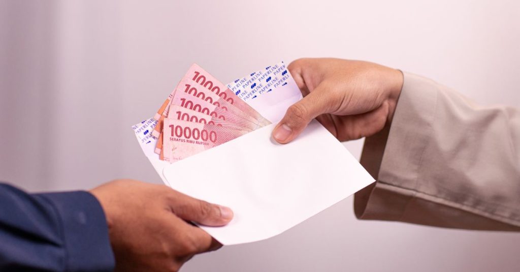 Tangan memberi dan menerima mata uang rupiah dalam amplop, melambangkan transaksi keuangan.