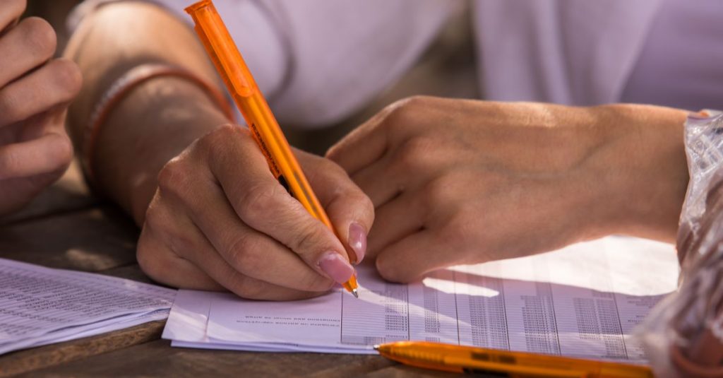 Bidikan jarak dekat tangan yang menulis pada dokumen dengan pena oranye, menyiratkan suasana bisnis atau pendidikan.