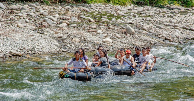 Sekelompok orang menikmati arung jeram di atas ban karet dengan tongkat sebagai kemudi.