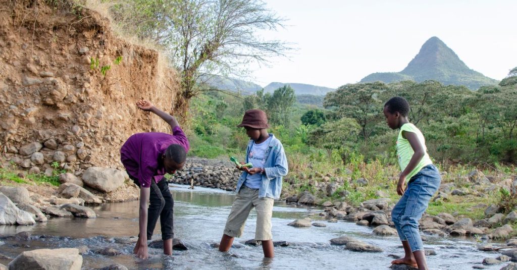 Tiga anak bermain di tepi sungai di lanskap pegunungan Afrika yang rimbun, menikmati alam.