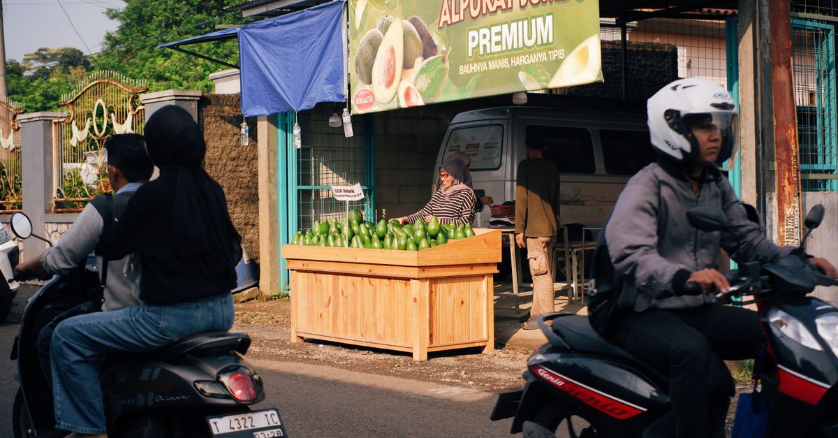 Pasar jalanan yang ramai di Jawa Barat dengan orang-orang yang mengendarai sepeda motor melewati kios alpukat.