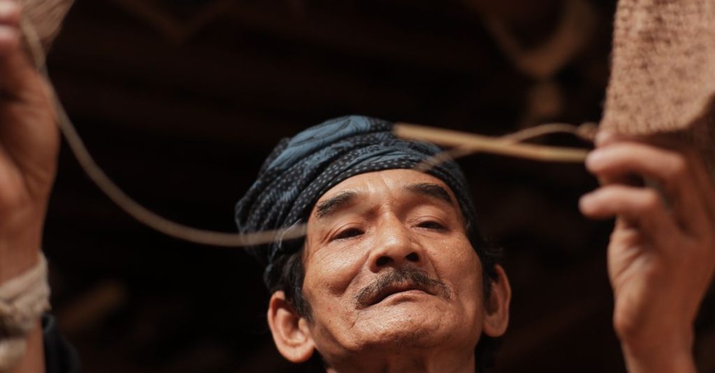 Gambar close-up seorang pengrajin lanjut usia Indonesia yang sedang menenun keranjang tradisional, menampilkan warisan budaya.
