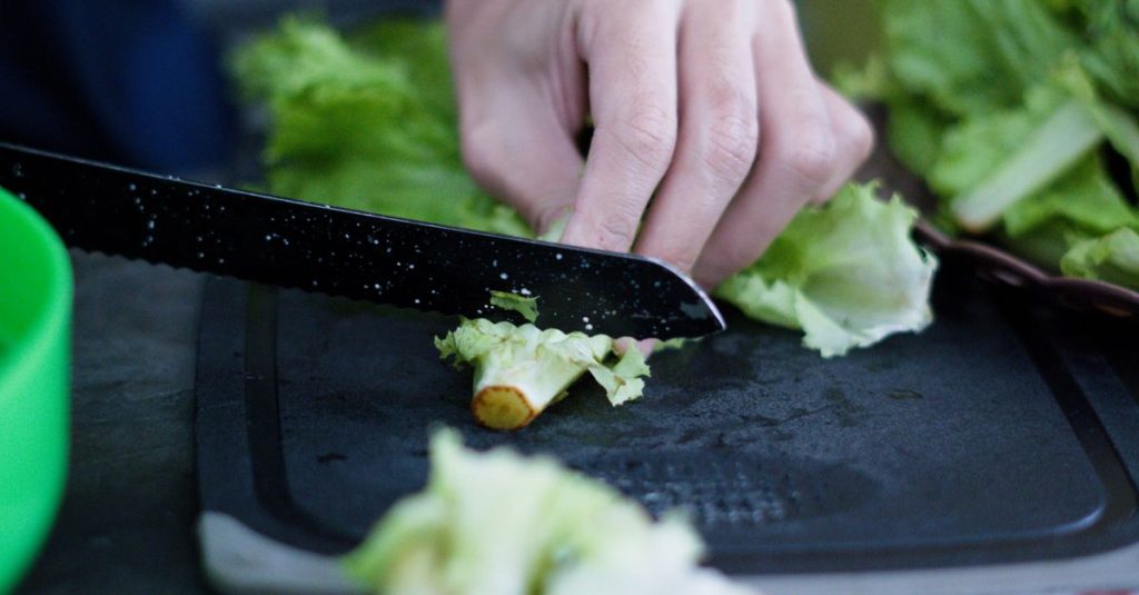 Close-up tangan yang mengiris selada segar di atas papan pemotong, sempurna untuk makanan sehat.