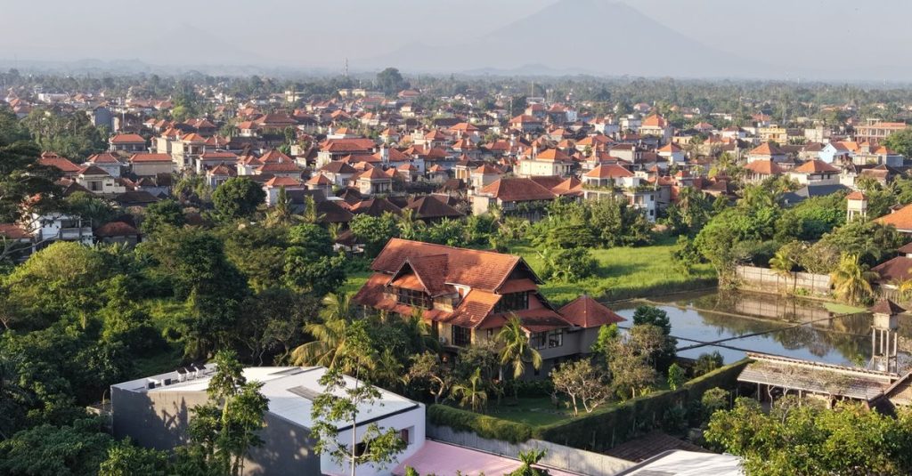 Pemandangan udara kota dengan rumah-rumah dan pepohonan hijau di bawah langit yang cerah.