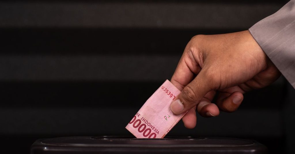 Gambar close-up sebuah tangan yang memasukkan uang kertas 100.000 Rupiah Indonesia ke dalam kotak donasi, menyoroti kegiatan amal dan memberi.