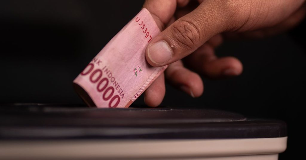 Gambar close-up sebuah tangan yang menyumbangkan uang kertas Rupiah Indonesia ke dalam kotak amal, melambangkan kemurahan hati.