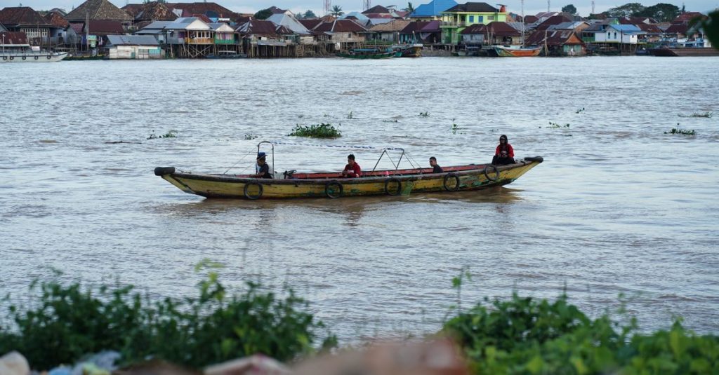 Pemandangan indah sebuah perahu tradisional di Sungai Musi di Palembang dengan orang-orang di dalamnya, dikelilingi oleh rumah-rumah di tepi sungai.