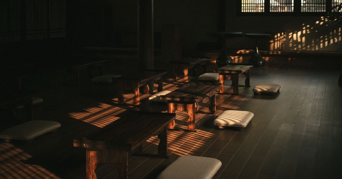 Ruang meditasi yang tenang dan diterangi cahaya matahari dengan meja kayu dan bantal di atas lantai kayu.