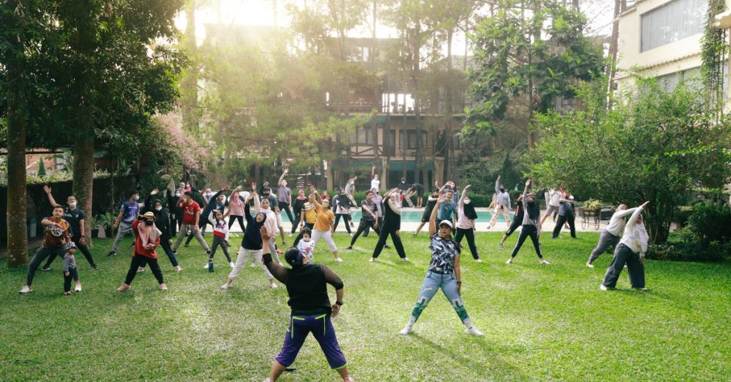 Sekelompok orang dengan latar belakang beragam berpartisipasi dalam kelas kebugaran di luar ruangan di taman yang cerah, mempromosikan kesehatan dan kesejahteraan.