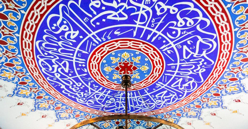 Kaligrafi Islam yang rumit menghiasi langit-langit berwarna-warni, menyoroti detail arsitektur yang unik.