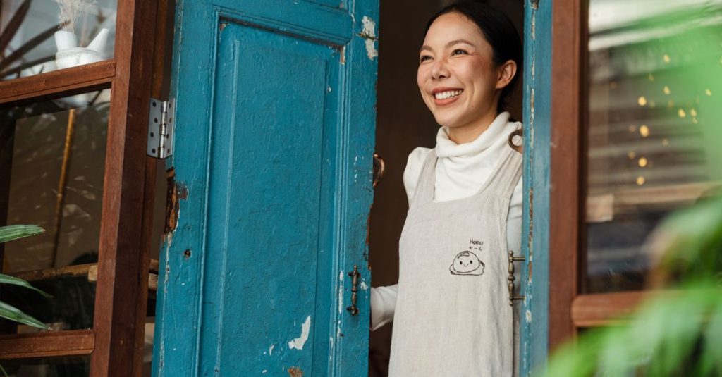 Wanita menawan bercirikan apron tersenyum bahagia di pintu kafe, menyambut tamu.