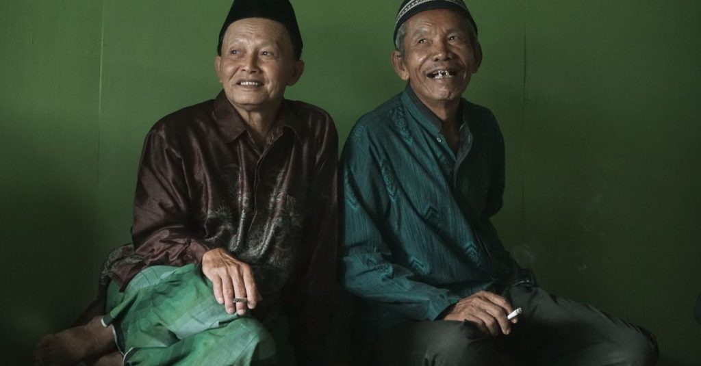Dua pria lansia dengan pakaian tradisional tersenyum hangat di dalam ruangan, melambangkan kebahagiaan.