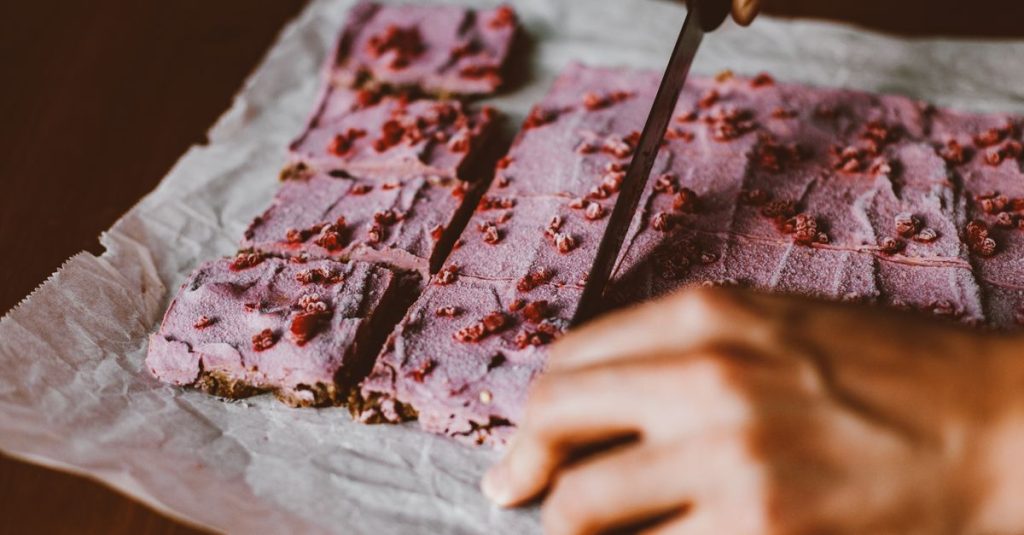 Kedekatan tangan yang sedang memotong batang pencuci mulut pink cerah dengan berry di atas kertas roti.