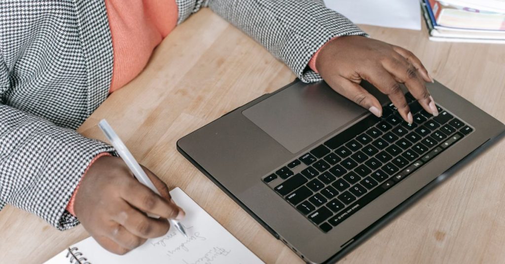 Wanita kulit hitam berpakaian resmi mengetik di laptop dan mencatat, menunjukkan keterampilan multitasking.