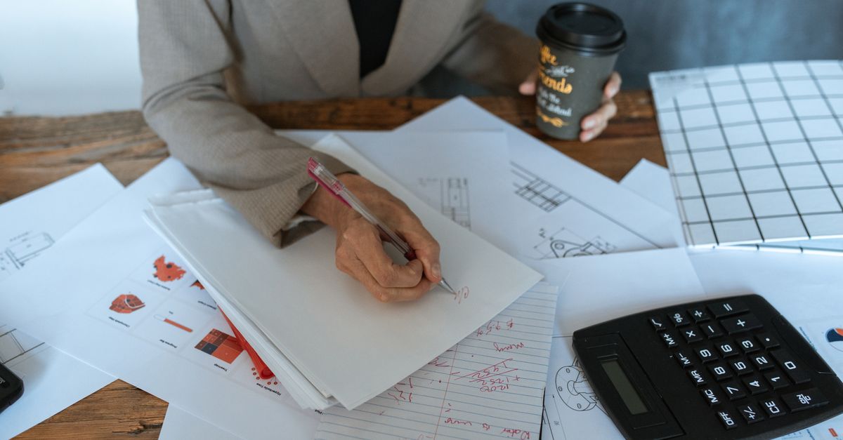 Close-up seseorang yang menulis di meja kantor dengan dokumen dan kopi, melambangkan produktivitas.