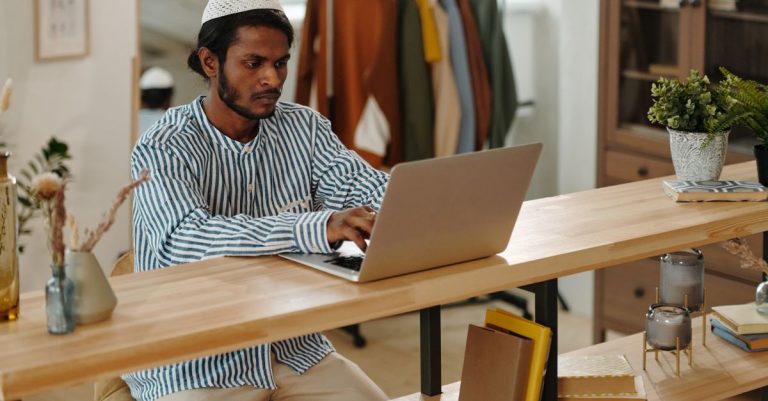 Pria muda dengan kemeja bergaris fokus pada laptop saat duduk di kantor rumah modern dengan dekorasi alami.