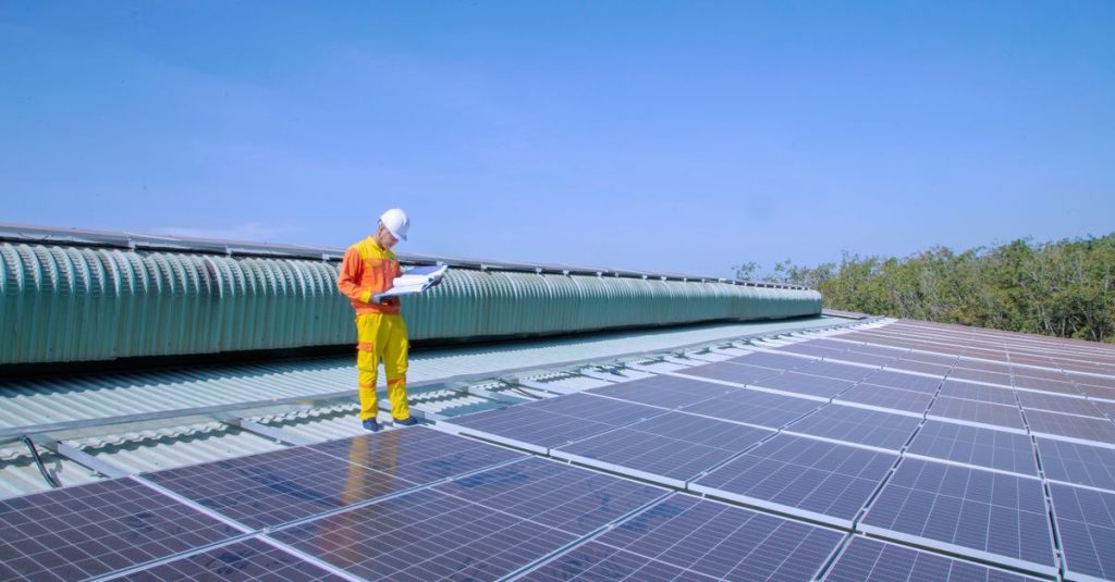 Seorang teknisi surya memeriksa panel surya di atap di bawah langit biru cerah, mempromosikan energi terbarukan.