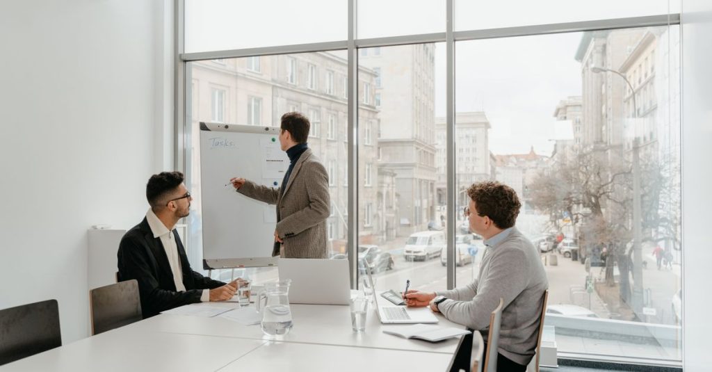 Tiga profesional terlibat dalam pertemuan bisnis di sebuah ruang konferensi yang cerah.