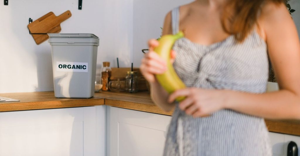 Seorang wanita di dapur memegang pisang dengan tempat sampah organik terlihat.