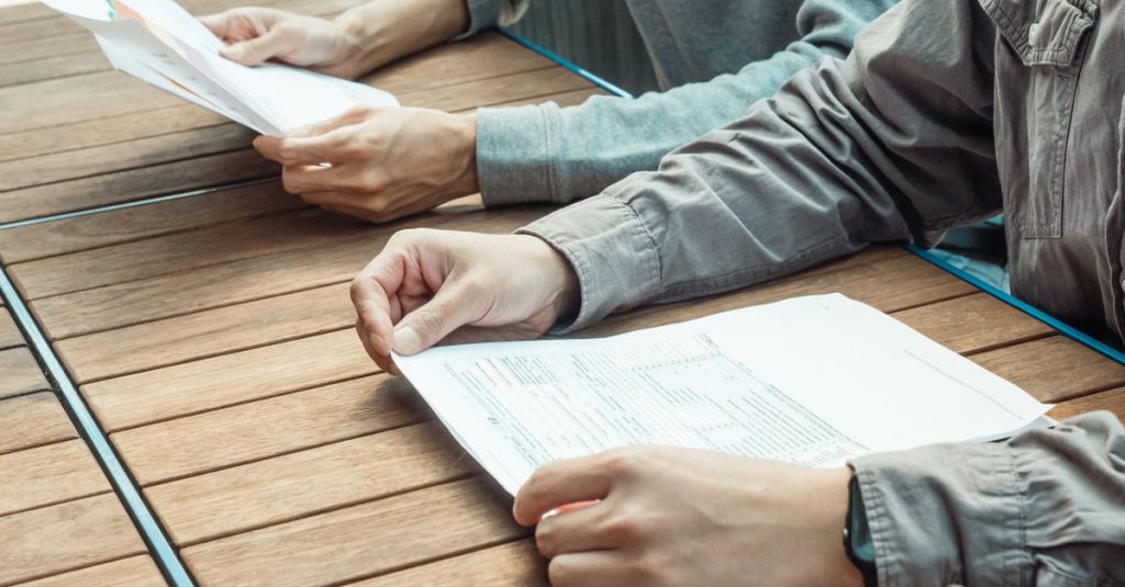 Dua orang sedang meninjau dokumen bisnis di meja luar ruangan, fokus pada pekerjaan kertas.