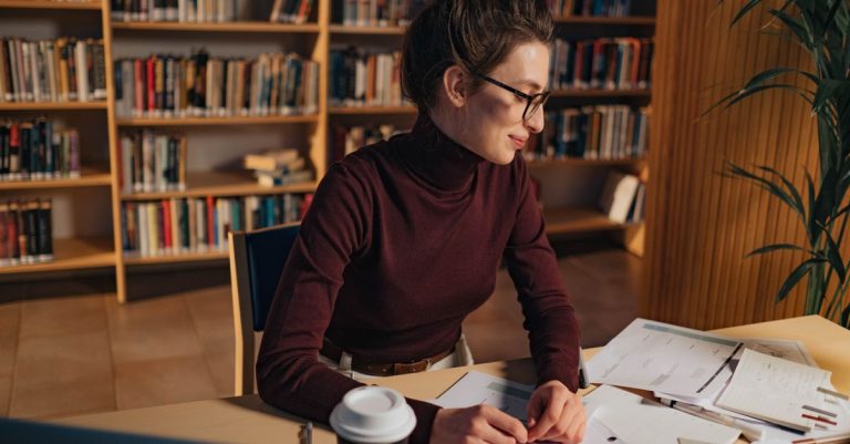 Seorang wanita di perpustakaan yang belajar dokumen dengan tekad, kacamata yang meningkatkan fokusnya.