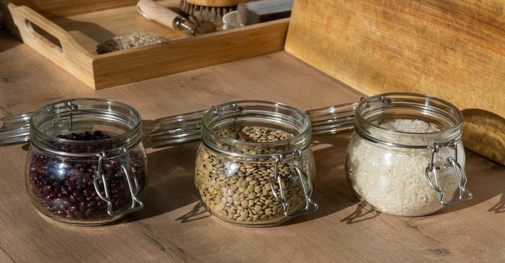 Stoples kaca berisi kacang, lentil, dan beras mentah di atas meja kayu dengan pencahayaan hangat.