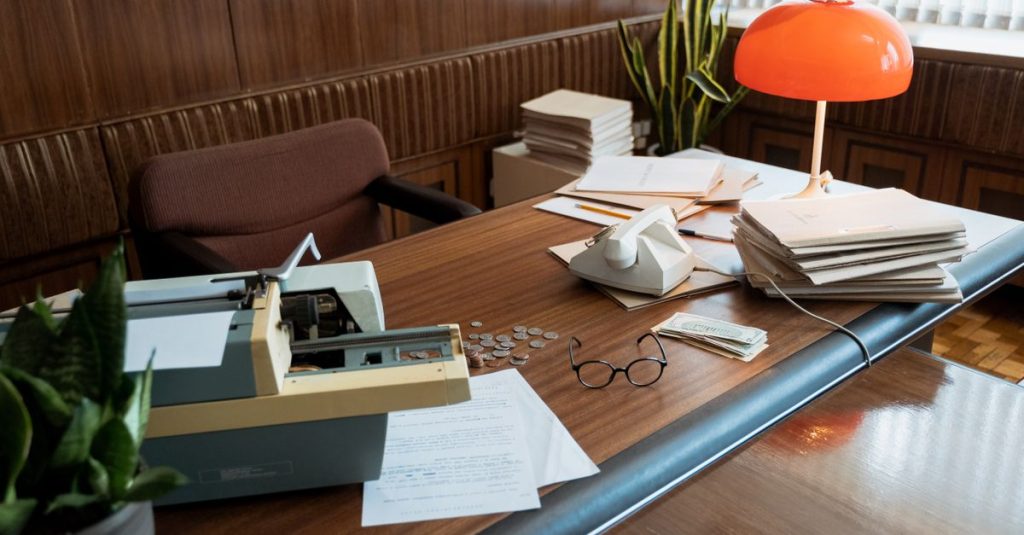 Meja kantor retro yang menampilkan mesin ketik, telepon vintage, dan tumpukan dokumen.