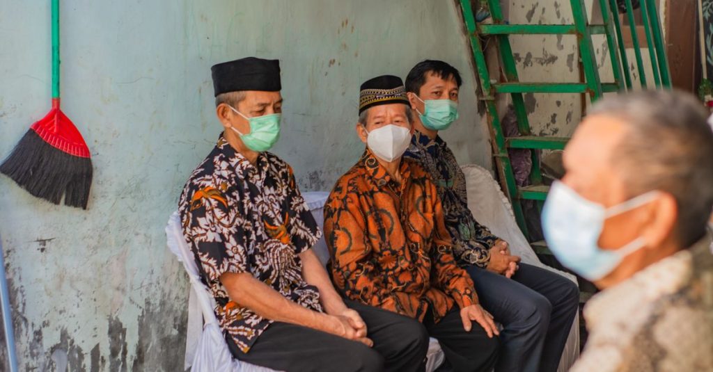 Tiga pria dengan pakaian tradisional mengenakan masker, duduk di dalam ruangan, menjaga protokol kesehatan.