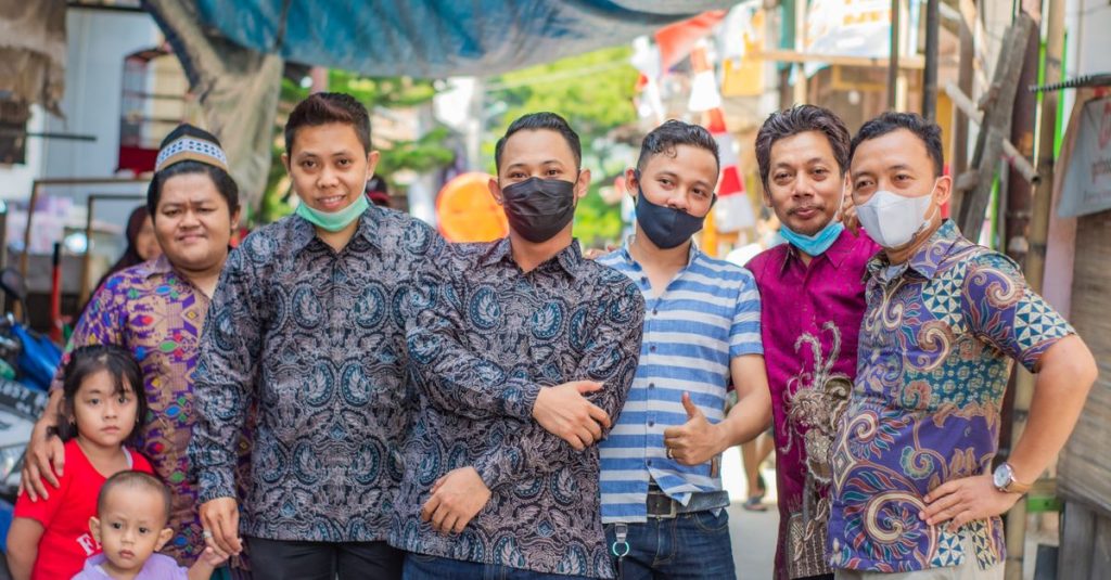 Sekelompok orang dewasa dan anak-anak yang beragam mengenakan masker wajah dan pakaian berwarna-warni, tersenyum bersama di luar ruangan.